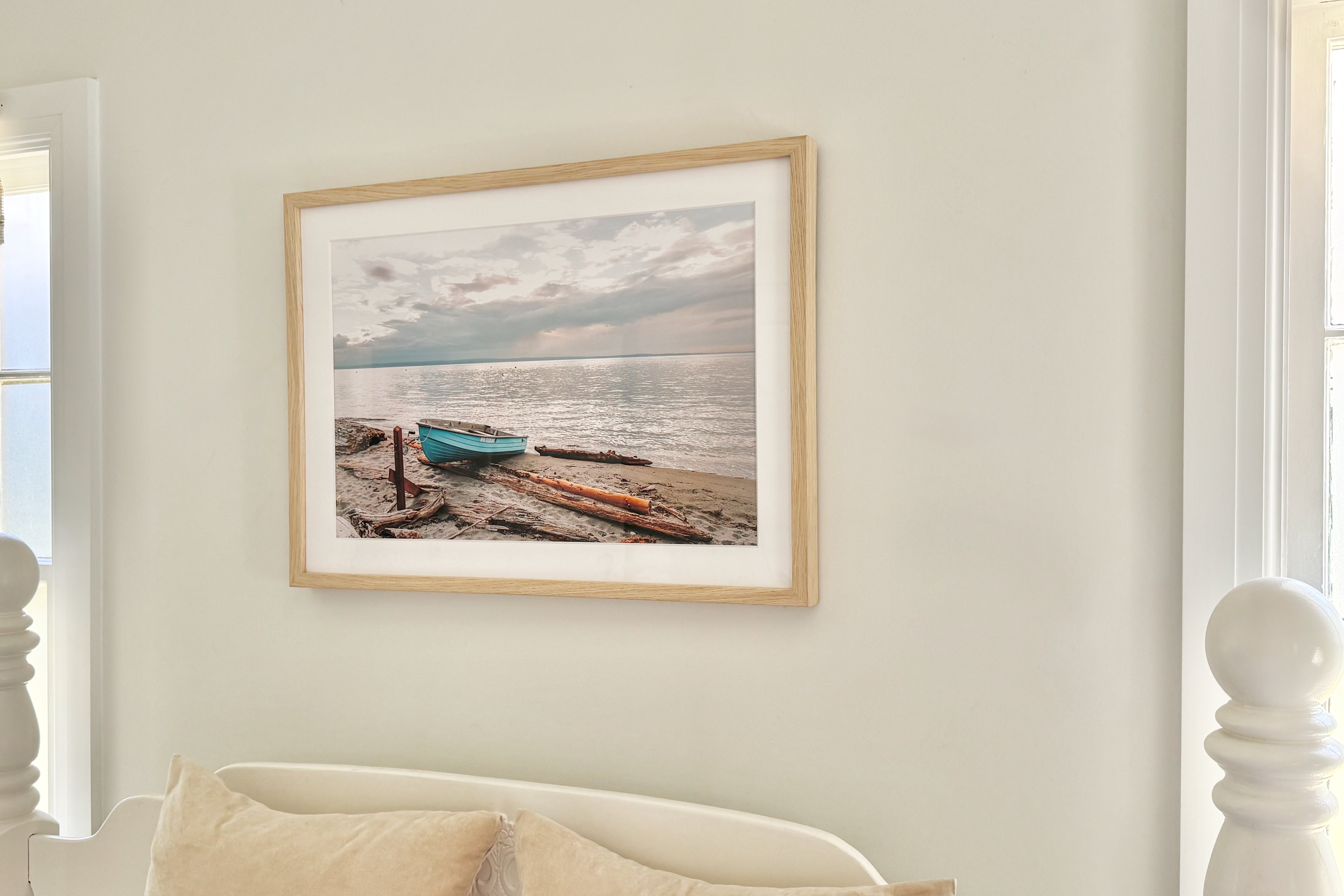 Framed photograph of a beach scene with boats on a white wall above a white bed by Jules Frazier.