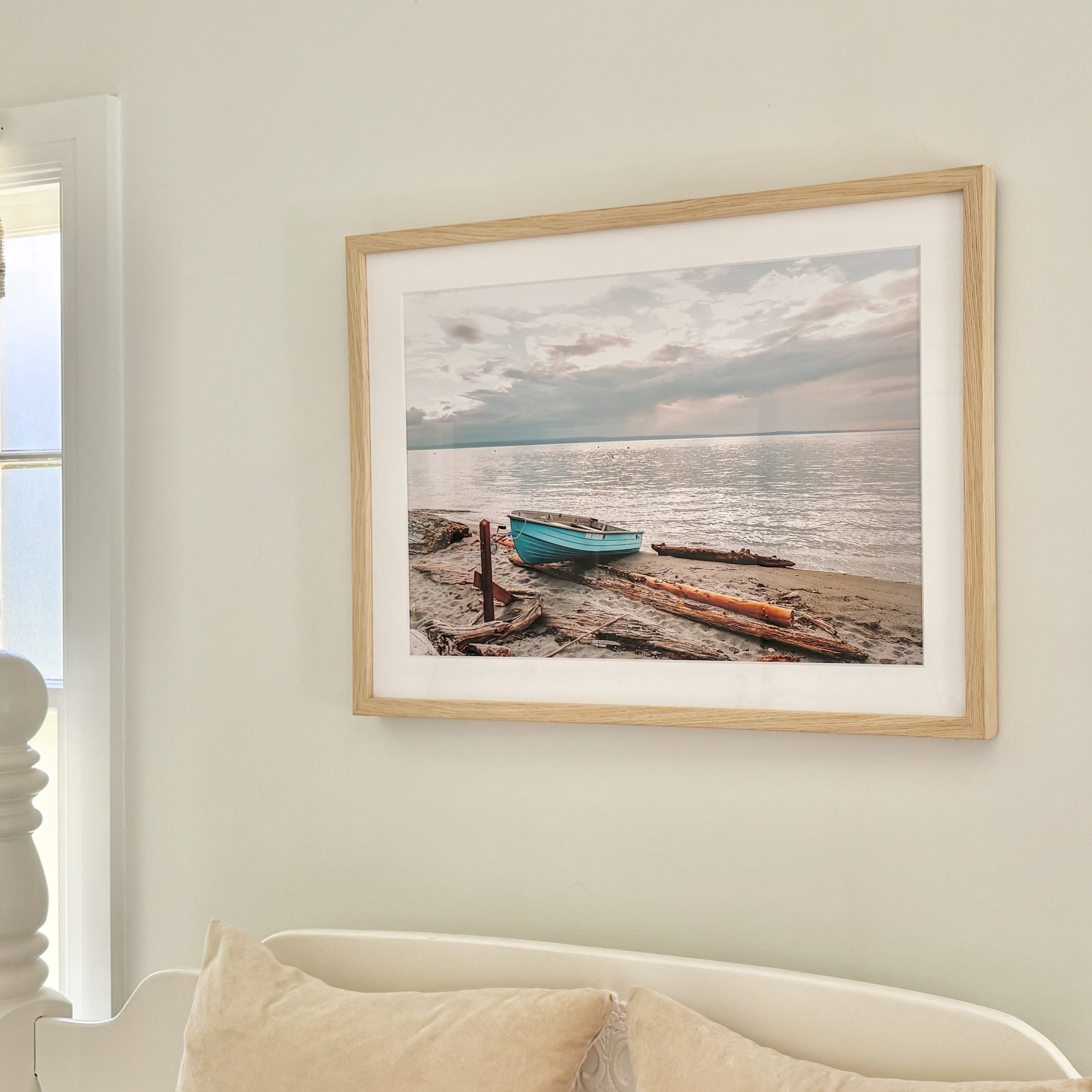 Framed photograph of a beach scene with boats on a white wall above a white bed by Jules Frazier.