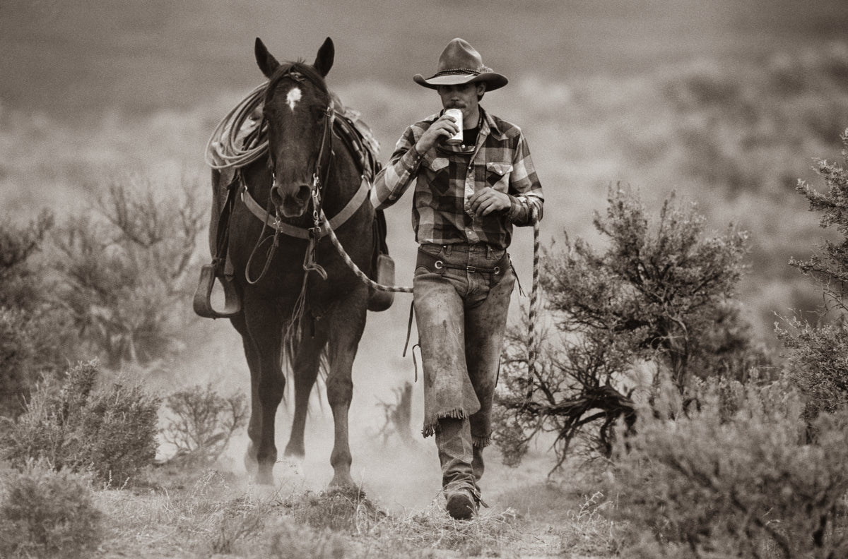 A black and white photograph of a cowboy in a wide-brimmed hat and plaid shirt walking alongside a horse through a dusty, brushy outdoor landscape.