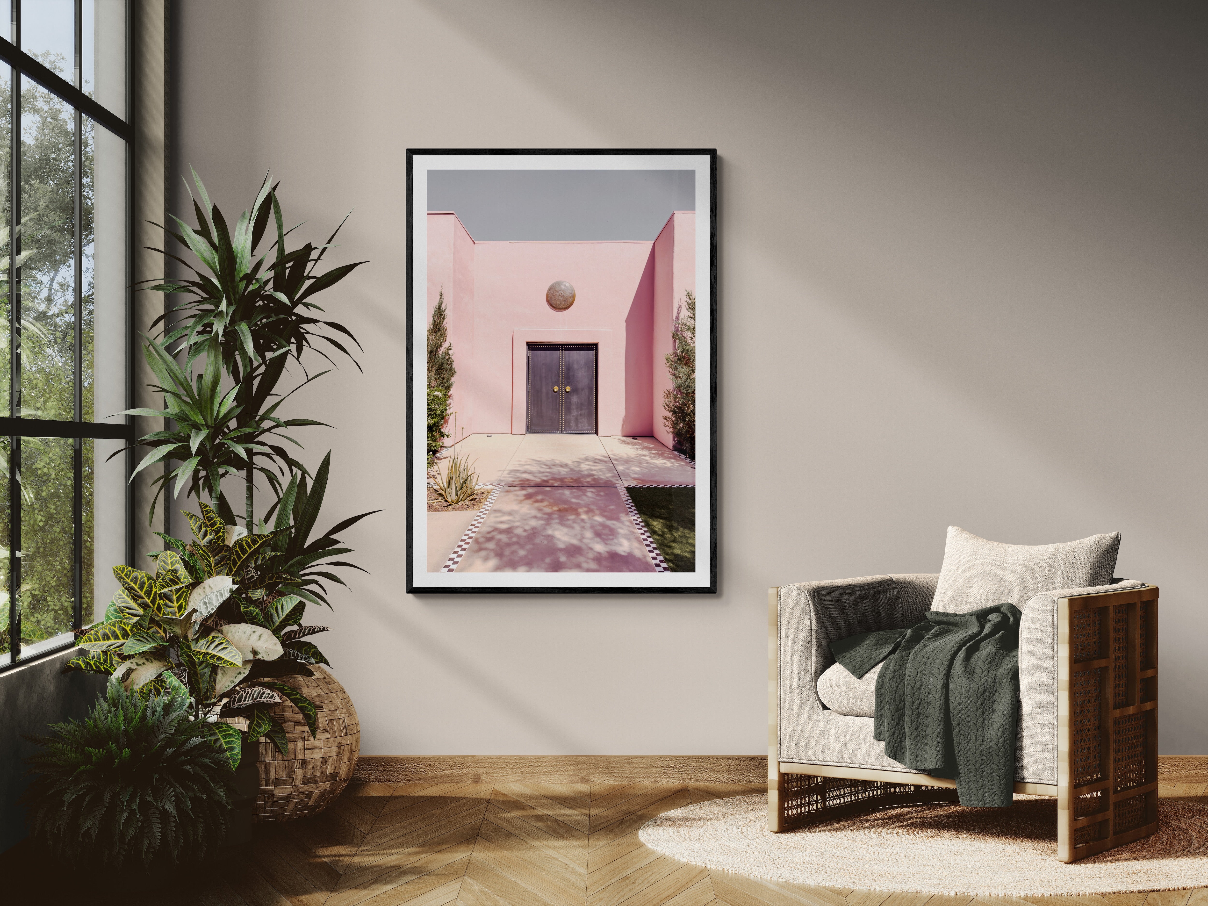 Framed artwork of a pink building on a wall in a room with a chair and plants.