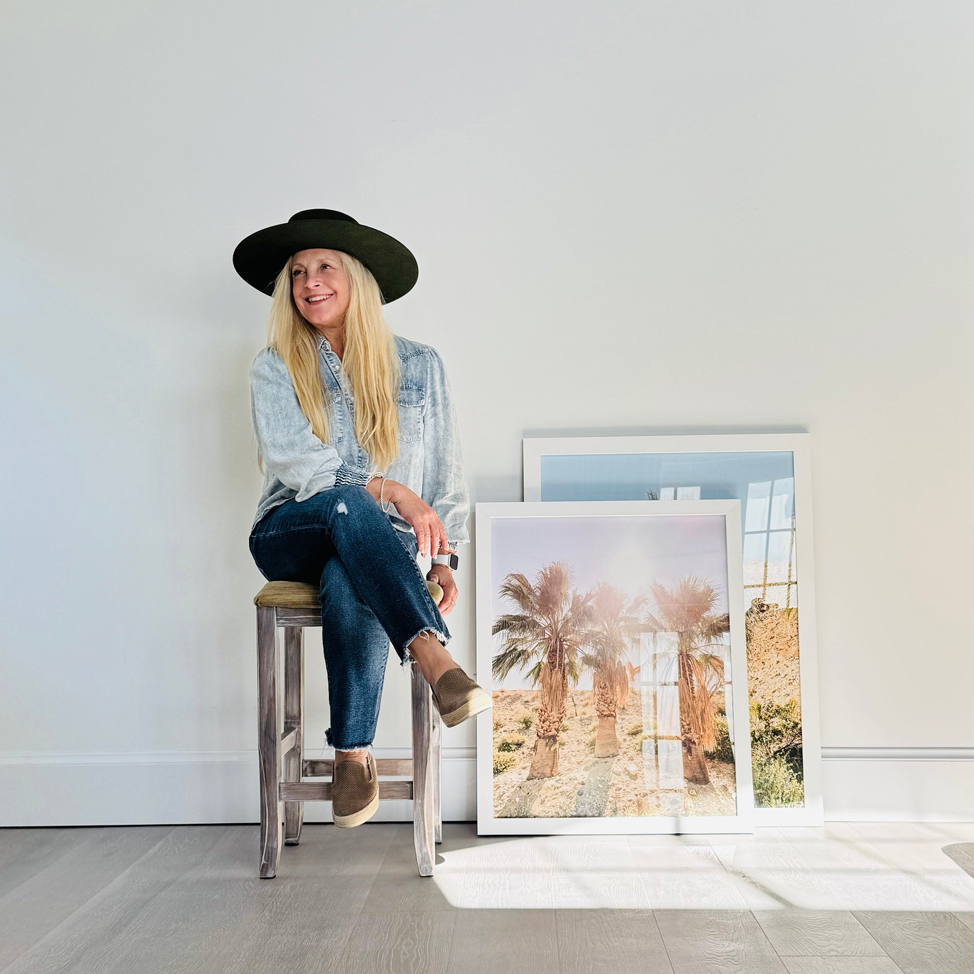 Jules Frazier sitting on a stool next to framed photographs of palm trees in a minimalistic room.