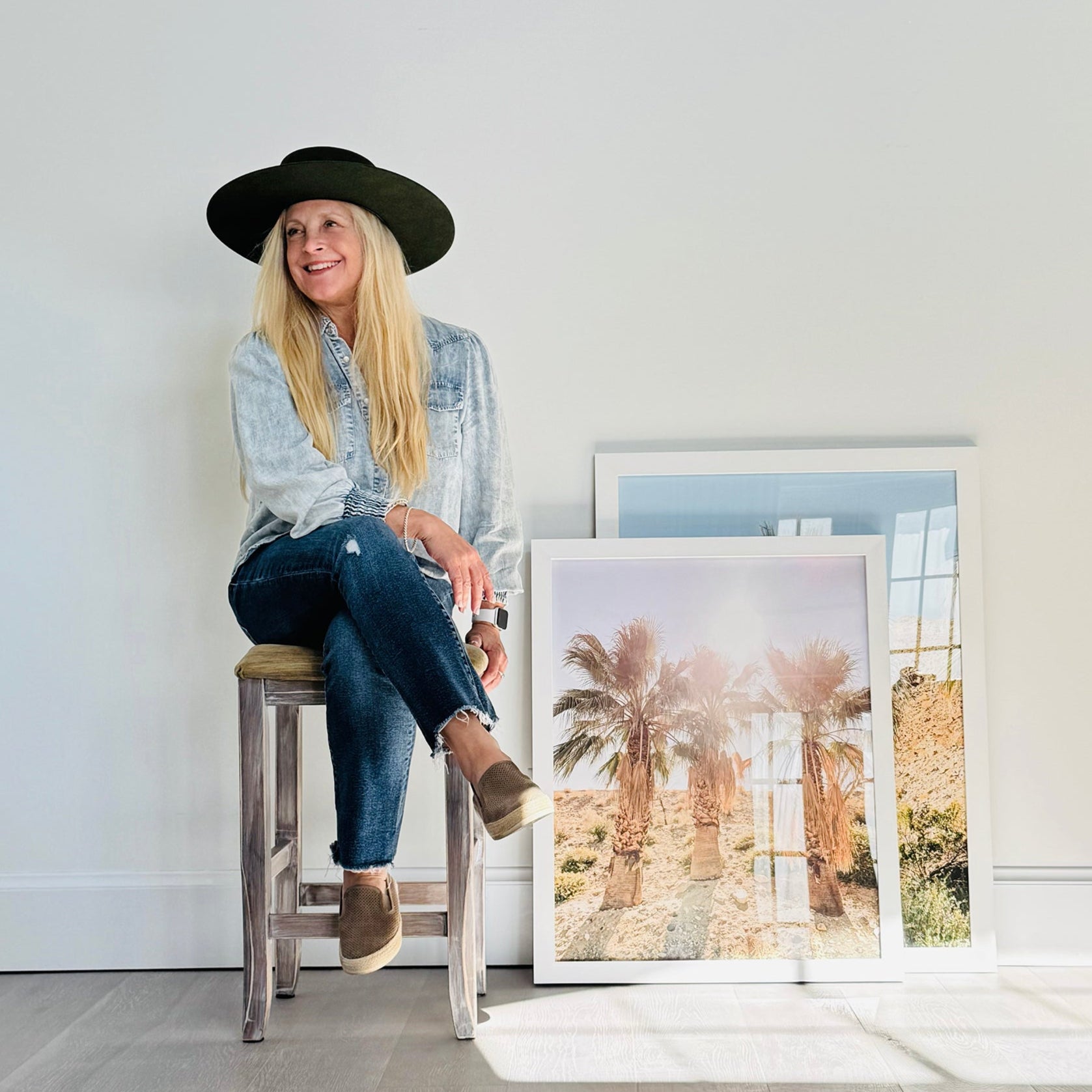 Jules Frazier sitting on a stool next to framed photographs of palm trees in a minimalistic room.