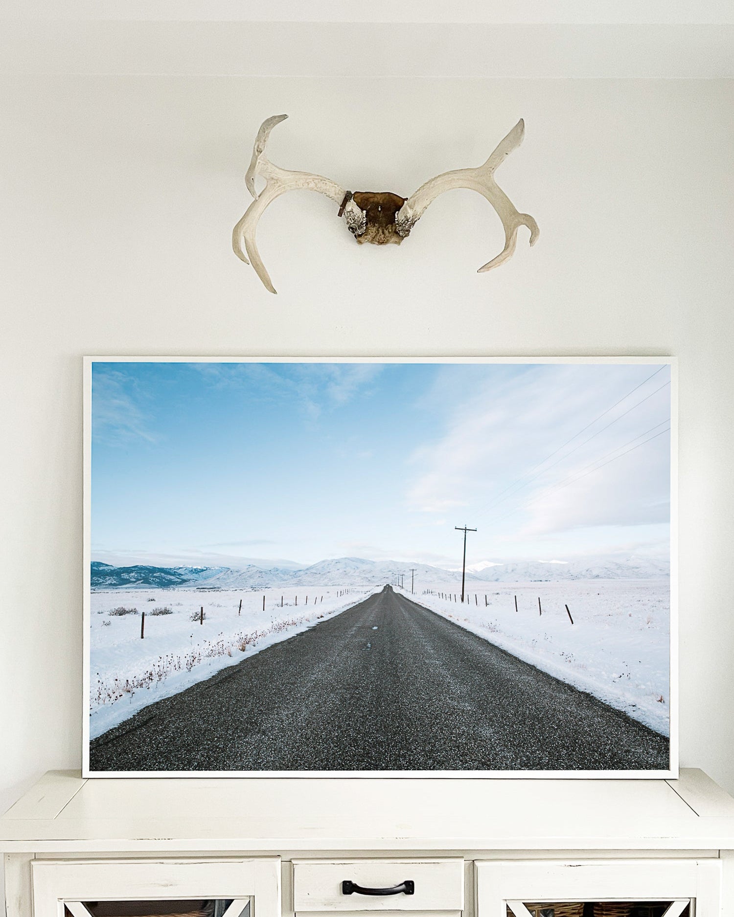 Framed picture of a snowy road with a deer antler above it on a wall.
