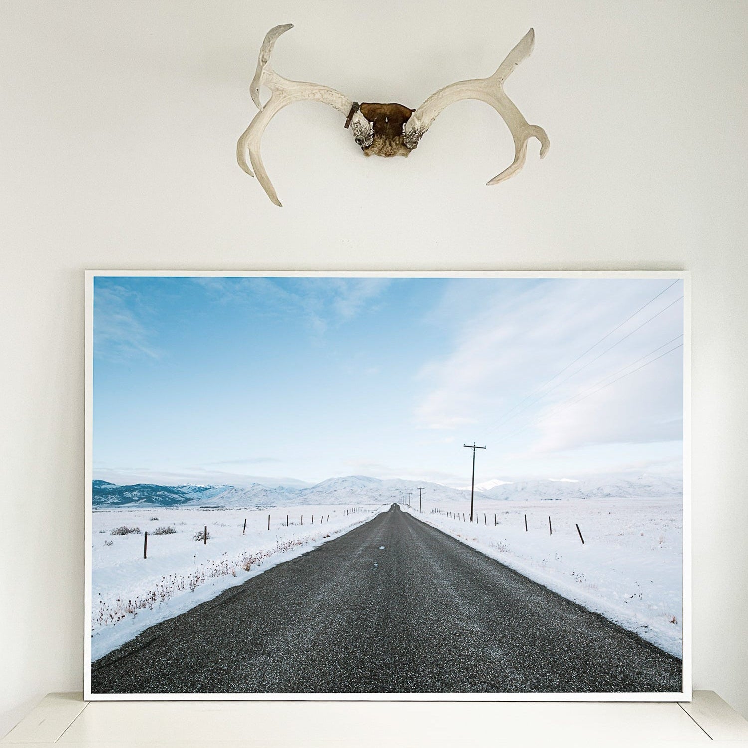 Framed picture of a snowy road with a deer antler above it on a wall.