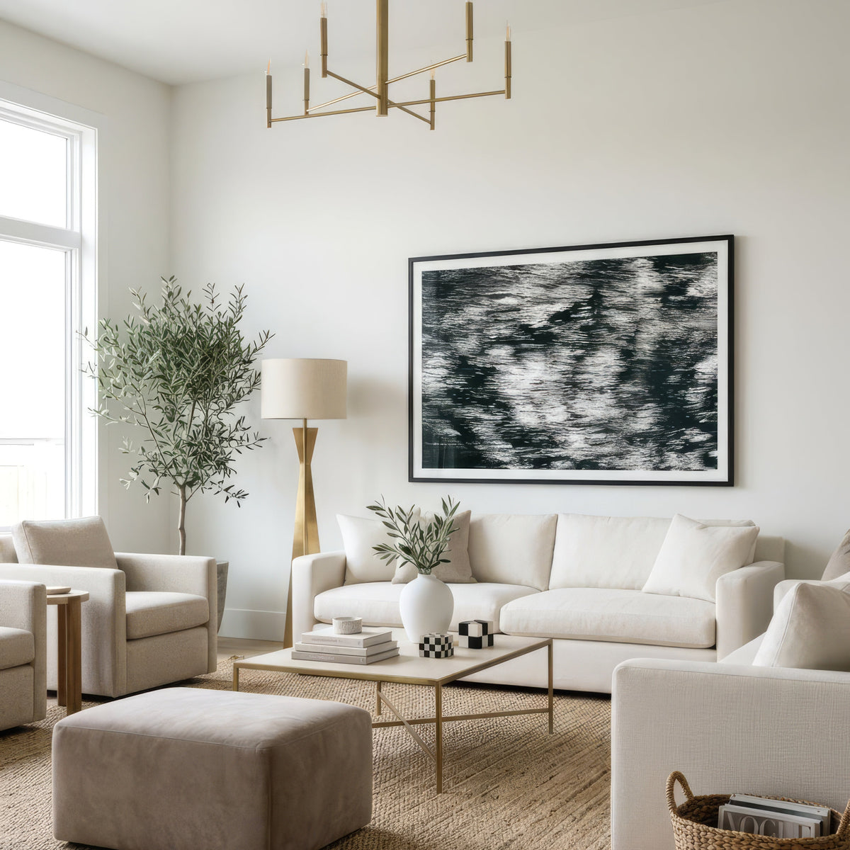 A modern living room featuring a white sectional sofa, a beige ottoman, a glass-top coffee table with gold legs, a large black and white abstract framed print on the wall, and a gold chandelier.