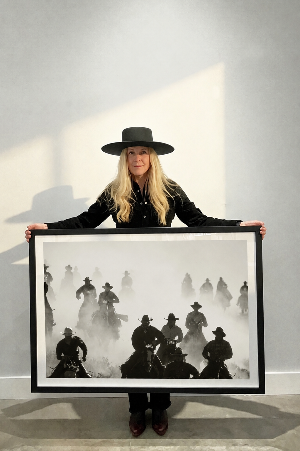 A woman wearing a black wide-brim hat and black outfit holds up a large framed black and white photograph depicting cowboys on horseback riding through heavy dust or fog.