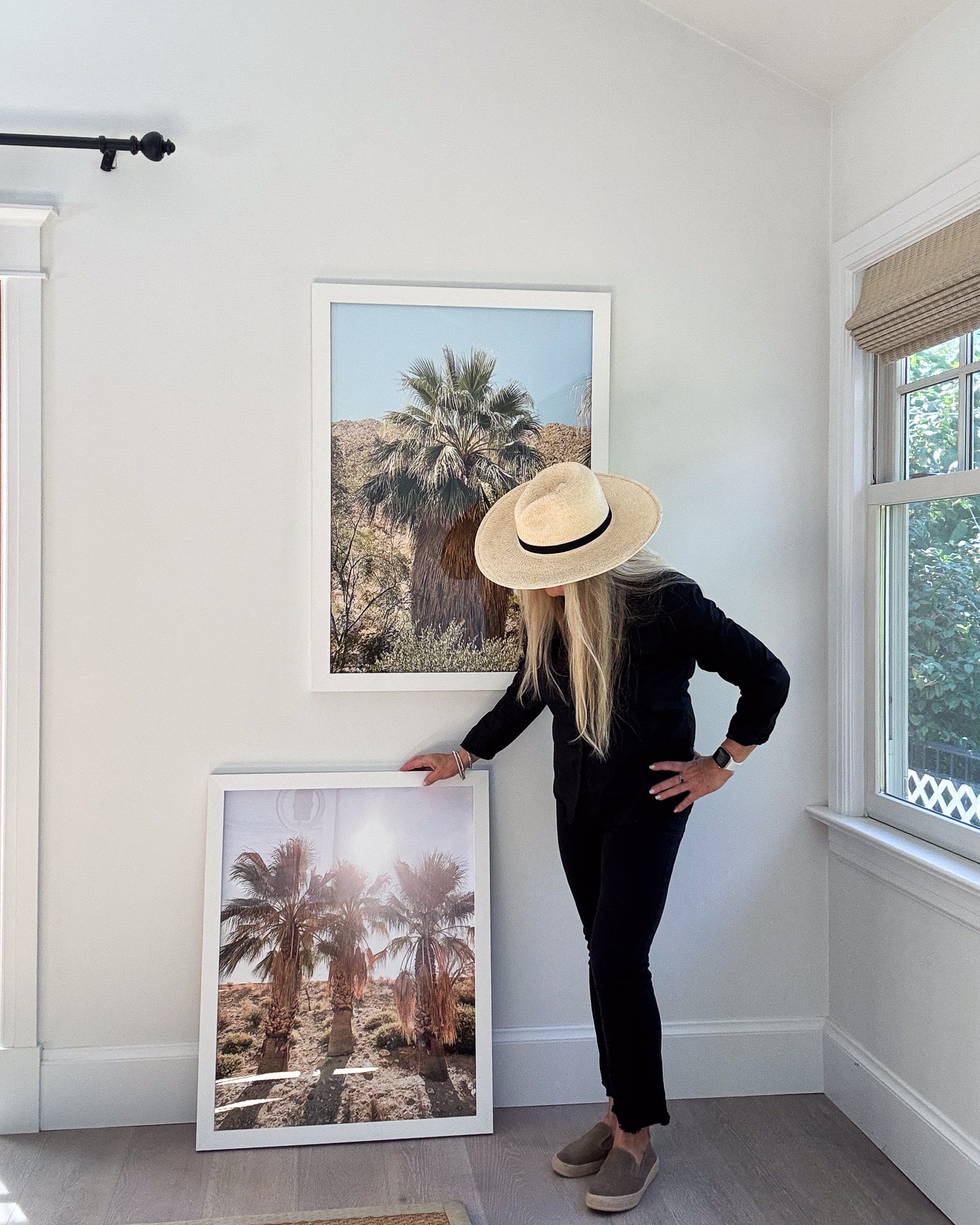 Person wearing a hat standing next to framed palm tree prints in a room with white walls and a window.