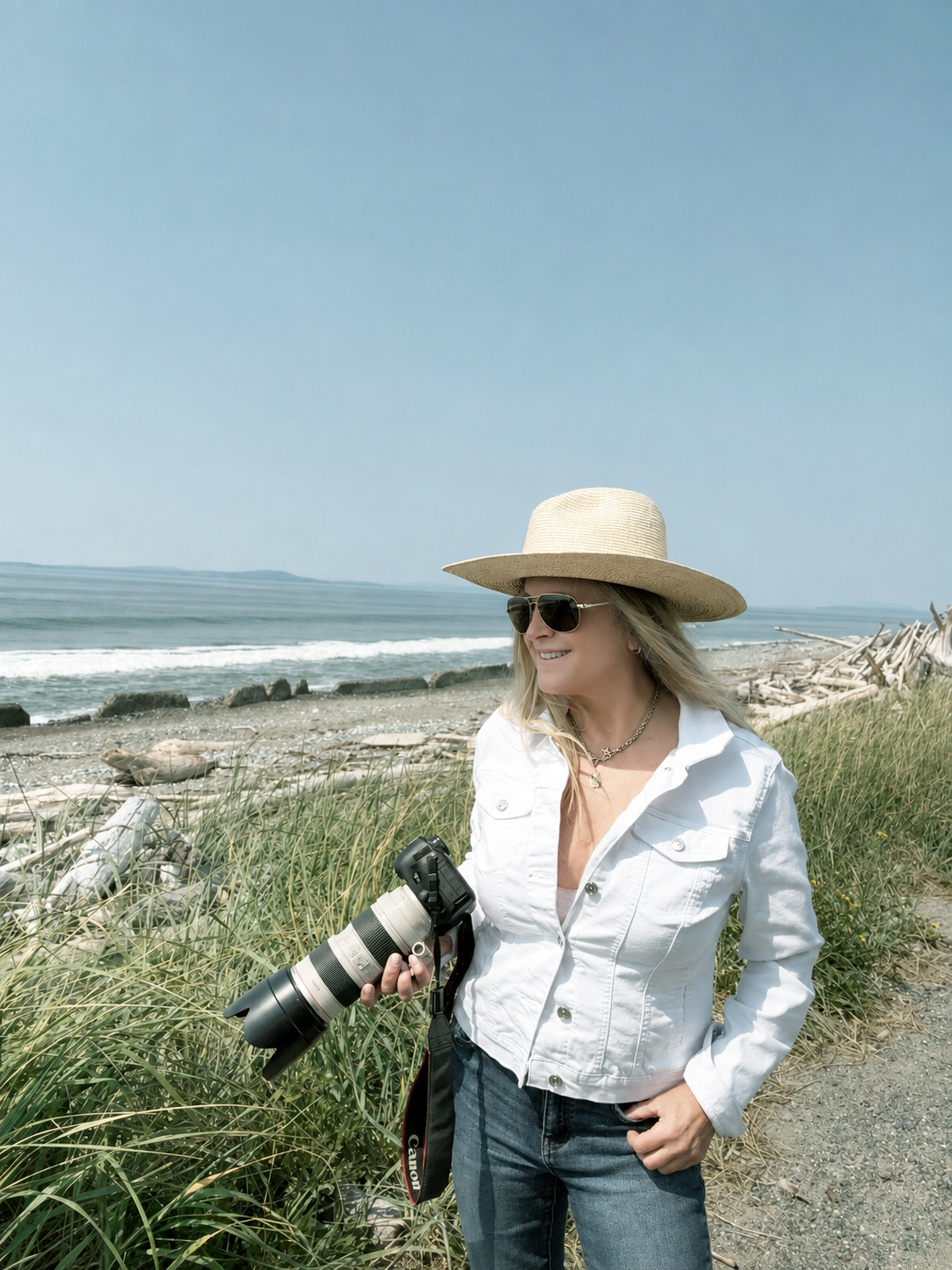 Jules Frazier standing on a beach path wearing a wide-brimmed straw hat, sunglasses, a white denim shirt, and blue jeans, holding a DSLR camera with a long lens. The background shows ocean waves, driftwood, and tall grass under a clear blue sky.