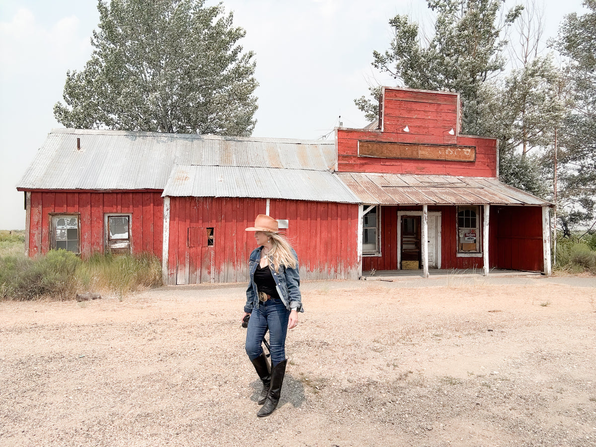 Person wearing a cowboy hat and boots walking in front of a red barn-like building.
