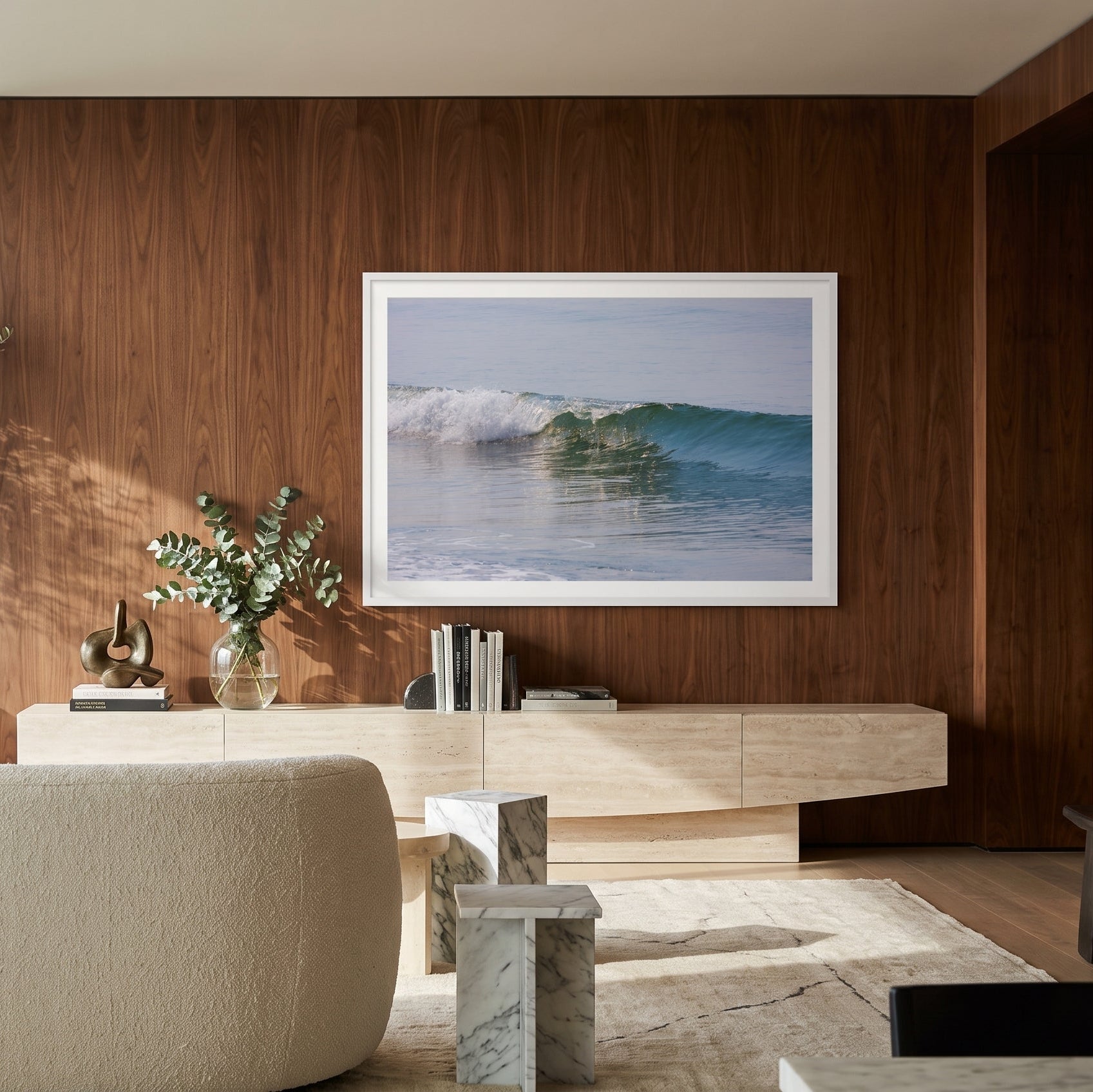A framed square art print depicting a blue ocean wave, displayed on a dark wood-paneled wall in a modern, sunlit living room with a beige sofa and minimalist furniture.