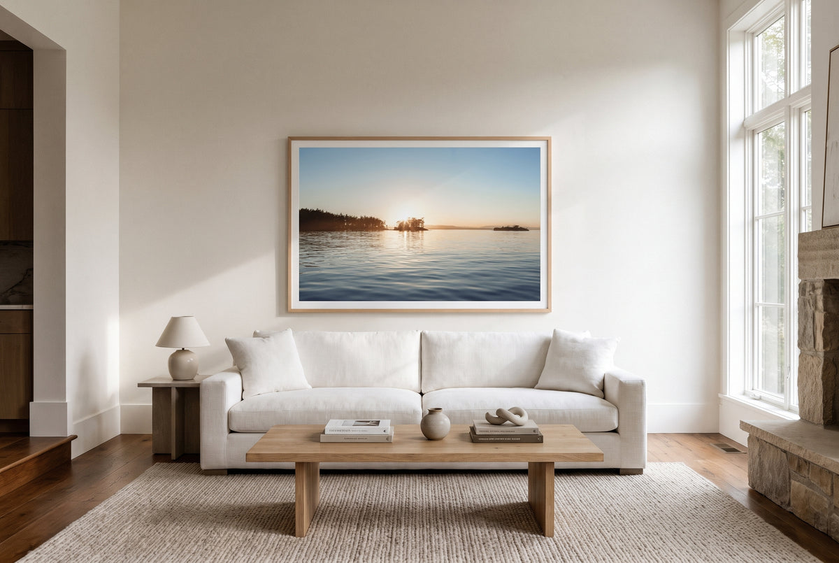A framed landscape art print of a calm lake at sunset hangs on a white wall above a modern white sofa in a bright, minimalist living room.